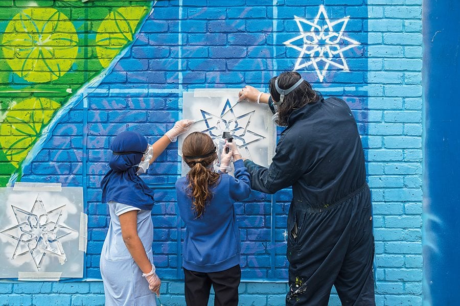 At Elmhurst School in Aylesbury, Teakster shows students how to use sticks to hold a stencil on the wall while spray painting the design. 